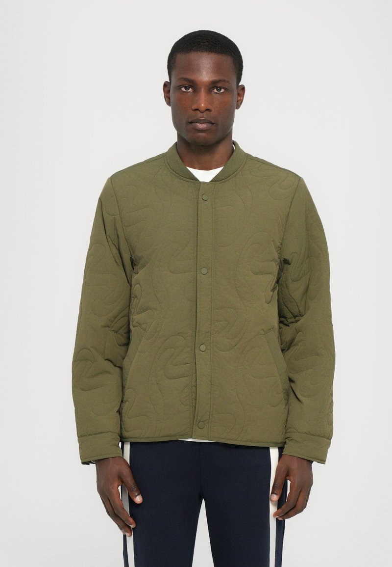 Jachetă bomber matlasată de culoare verde olive, cu guler rotund, nasturi prin snap pe față și un model texturat subtil. Modelul poartă un tricou alb dedesubt.
