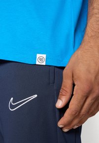 Blå bomulds T-shirt med en hvid etiket, der siger "90" ved kanten, parret med mørkeblå bukser med et hvidt broderet Nike-logo.