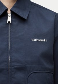 Μπλε μπουφάν με φερμουάρ και γιακά, με λευκό κεντημένο λογότυπο «carhartt» στο αριστερό στήθος, που φοριέται από ένα άτομο.