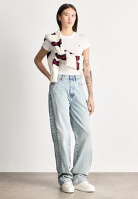 Jauns cilvēks ar tetovējumiem valkā baltu T-kreklu, gaisīgi zilas platām biksēm, baltas sporta kurpes un pār pleciem pārmetušu svītrainu džemperi.