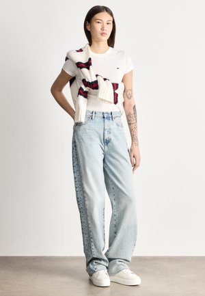 Jauns cilvēks ar tetovējumiem valkā baltu T-kreklu, gaisīgi zilas platām biksēm, baltas sporta kurpes un pār pleciem pārmetušu svītrainu džemperi.