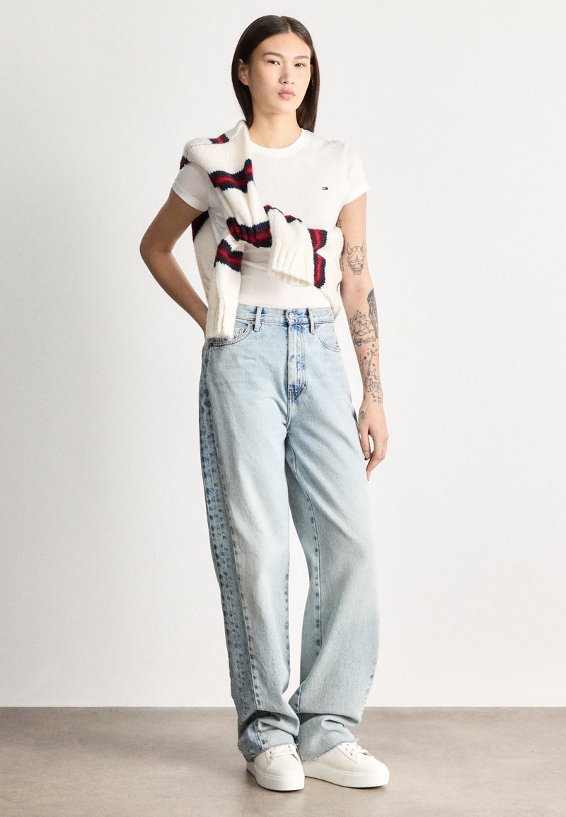 Jauns cilvēks ar tetovējumiem valkā baltu T-kreklu, gaisīgi zilas platām biksēm, baltas sporta kurpes un pār pleciem pārmetušu svītrainu džemperi.