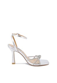 Sandalo con tacco alto argento, punta quadrata, cinturini incrociati e dettaglio a nodo decorativo. Presenta una sottile cinghia per la caviglia e una finitura metallica.
