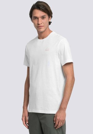 Giovane uomo con capelli castani di media lunghezza che indossa una t-shirt bianca semplice a maniche corte e pantaloni scuri, in piedi su uno sfondo chiaro.