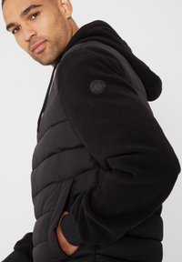 Sort polstret jakke med hætte foret med fleece, der har et gummi logo badge på skulderen og sidelommer. Glat tekstur og varmt design.