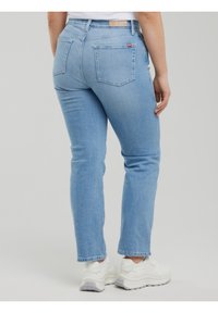 Isik kandmas helesiniseid teksapükse ja valgeid tossusid, nähtud tagantpoolt ühtlase hele tausta taustal.