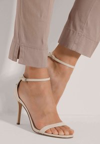 Beige high heelsandalen met dunne banden en een enkelgesp, gecombineerd met lichtgekleurde, cropped broeken met zijslits.