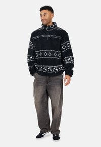 Pullover in pile nero con motivi geometrici bianchi, colletto a metà zip e polsini a coste, abbinato a jeans grigi sbiaditi e sneakers nere.
