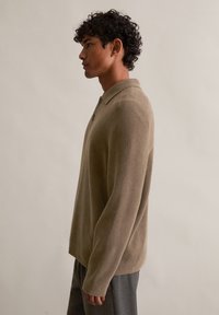 Beżowa, długorękawowa koszulka polo z teksturowanej tkaniny, z miękkim kołnierzykiem i dwoma guzikami, w zestawie z szarymi spodniami.