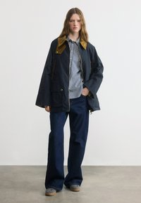 Νεαρή γυναίκα που φοράει μπλε σκούρο μπουφάν με καφέ γιακά, μπλε ριγέ πουκάμισο, τζιν παντελόνι με φαρδιά μπατζάκια και γκρι αθλητικά παπούτσια, στέκεται σε τσιμεντένιο δάπεδο.