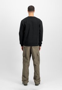 Uomo in piedi di spalle, indossa una felpa nera, pantaloni cargo verde oliva con tasche e scarpe nere, su uno sfondo bianco uniforme.