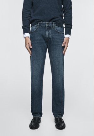 Man draagt donkerblauwe jeans, zwarte leren schoenen en een navy trui met zichtbare witte overhemdsmouwen, staand tegen een effen achtergrond.