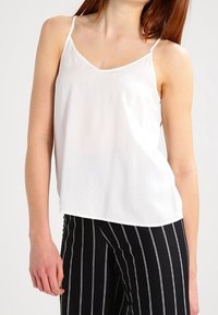 Mujer vestida con una blusa sin mangas blanca y pantalones negros con rayas verticales blancas, de pie frente a un fondo liso.