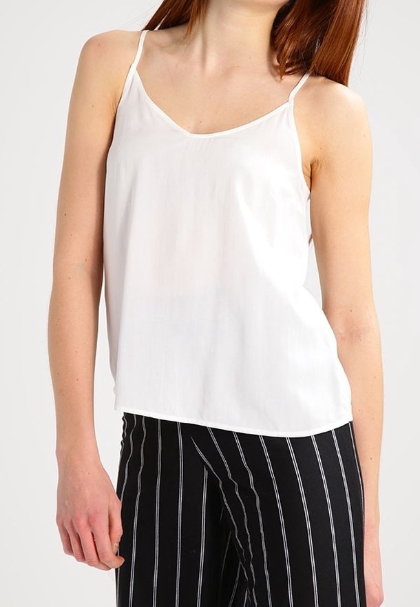 Mujer vestida con una blusa sin mangas blanca y pantalones negros con rayas verticales blancas, de pie frente a un fondo liso.