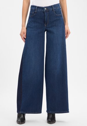 Wijde donkerblauwe denim jeans gedragen met zwarte enkellaarzen, zichtbaar van taille tot voeten tegen een witte achtergrond.