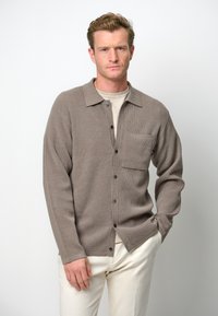 Brun ribbad cardigan med krage, knappfront och en enda bröstficka, bärs över en ljus t-shirt och matchas med vita byxor.