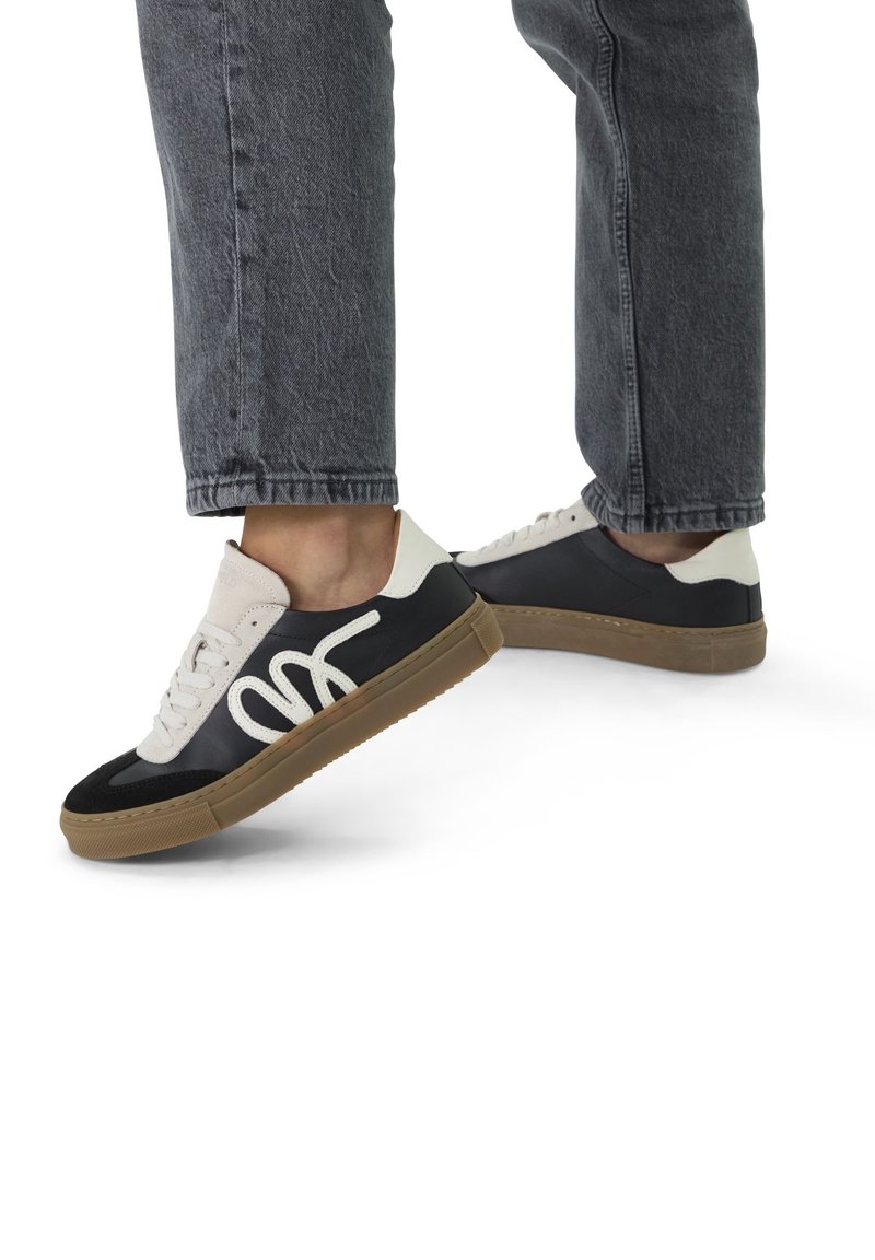 Pieds portant des baskets noires et blanches avec des semelles marron et des lacets blancs, assortis à un jean gris foncé sur fond blanc.