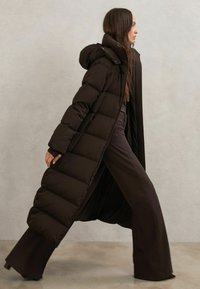 Cappotto imbottito lungo nero con cappuccio, caratterizzato da sezioni trapuntate e vestibilità ampia, abbinato a pantaloni neri a gamba larga. Tessuto liscio.