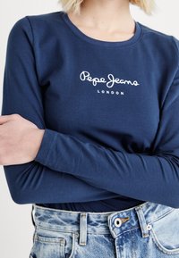 Mörkblå långärmad t-shirt med vit "Pepe Jeans London" logotyp, gjord av mjuk bomull, figursydd design, tillsammans med ljusblå slitna denimjeans.