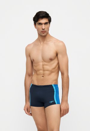 Young man with muscular build wearing navy blue and light blue swim trunks standing against a plain white background.