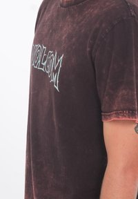 Mies, joka on pukeutunut haalistuneeseen tummanpunaiseen T-paitaan, jossa rintaosassa on sinisellä tyylitelty teksti "VOLCOM". Paidasta näkyy osa tatuoidusta käsivarresta.