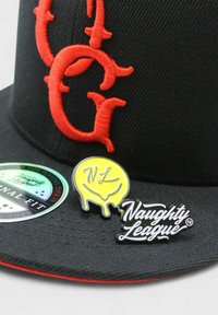 Mustapää, jossa on punaiset brodeeratut kirjaimet. Kaksi nappia: keltainen valuvan muotoilun, jossa lukee "NL" ja musta nappi, jossa on teksti "Naughty League." Kerroksellisia tarroja.