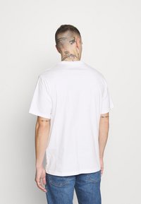 Bílá bavlněná tričko s krátkými rukávy, kulatým výstřihem a rovného střihu. Má minimální branding na lemu.