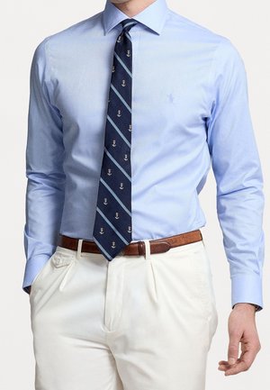 Mann trägt hellblaues Hemd mit langen Ärmeln, dunkelblaue gestreifte Krawatte mit Anker-Mustern, braunen Gürtel und weiße Hose mit Händen in den Taschen.