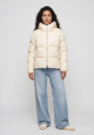 Frau trägt eine cremefarbene Daunenjacke, hellblaue Jeans mit weitem Bein und weiße Sneakers, steht vor einem einfarbigen hellen Hintergrund.
