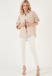 Camicia oversize beige con bottoni e maniche arrotolate, abbinata a jeans skinny bianchi e décolleté beige. Tessuto morbido con una vestibilità rilassata.