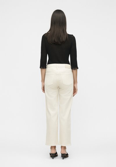 Γυναίκα με ίσια σκουρόχρωμα μαλλιά που φορά μαύρο τοπ με μανίκια τριών τετάρτων, λευκό παντελόνι με ψηλή μέση και ίσια γραμμή, και μαύρα ψηλοτάκουνα σανδάλια, όρθια.