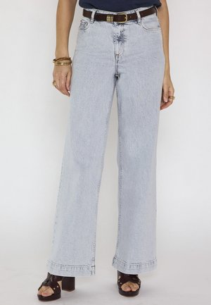 Personne portant un jean large taille haute, délavé clair, avec une ceinture marron, des sandales à talons foncées et des bracelets et bagues dorés sur fond blanc.