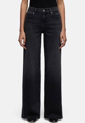 Donna che indossa jeans neri a vita alta e gamba larga e scarpe nere, in piedi con le mani rilassate lungo i fianchi su sfondo bianco.