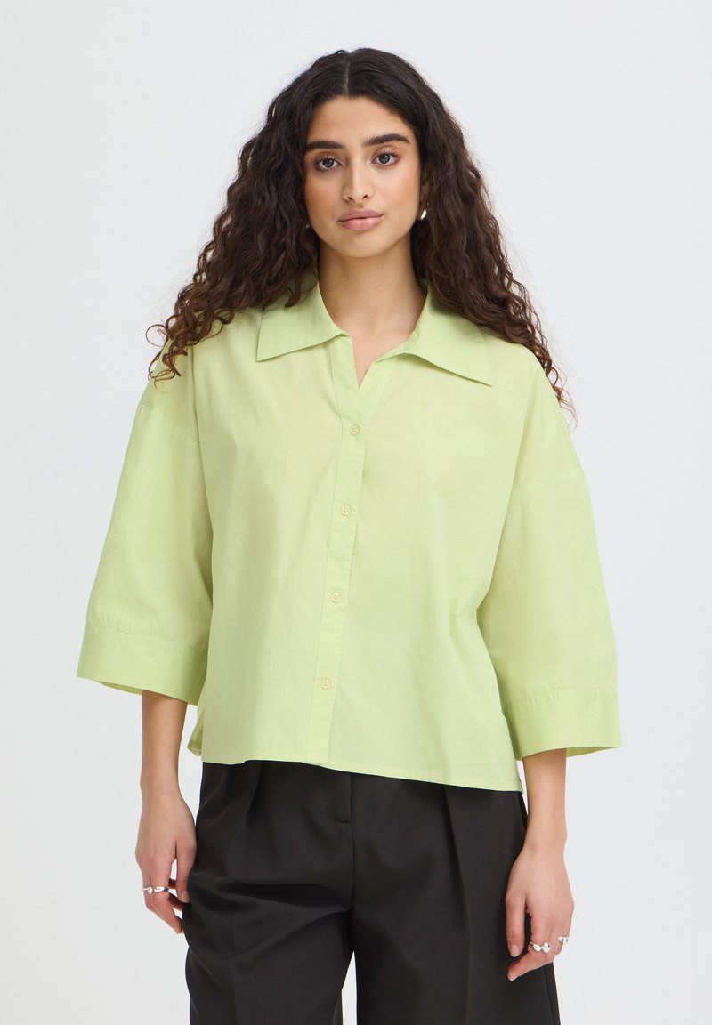 Femme aux longs cheveux bouclés porte une chemise verte claire boutonnée avec un large col et un pantalon noir, debout devant un fond uni.
