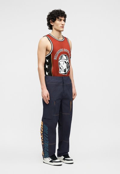 Άνδρας που φοράει κόκκινη μπλούζα μπάσκετ χωρίς μανίκια με γραφικό αστροναύτη, μπλε ναυτικού χρώματος παντελόνι cargo με το κείμενο "Billionaire Boys Club" και μαύρα και άσπρα αθλητικά παπούτσια.