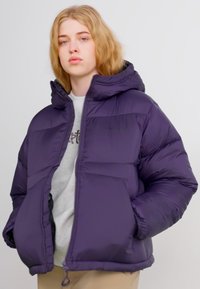 Ljubičasta jakna s kapuco, s prednjim zadrgo, prešito zasnovo in dvema stranskima žepoma. Narejena iz gladkega, izoliranega materiala.