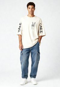 Beige ylisuurten t-paitojen grafiikalla, mustalla tekstillä "TAKE CARE" ja "LOVE" hihoissa. Yhdistetty vaaleansinisiin cargo-farkkuihin ja valkoisiin lenkkareihin.