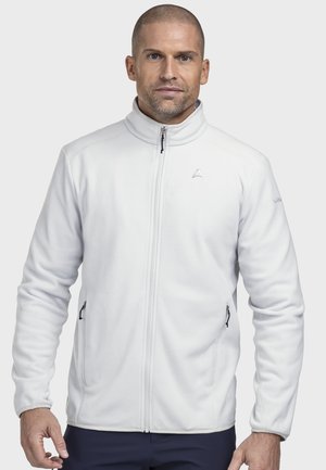 Mann mit kurzen Haaren, der eine weiße Fleecejacke mit Reißverschluss und Seitentaschen trägt und vor einem schlichten Hintergrund steht.