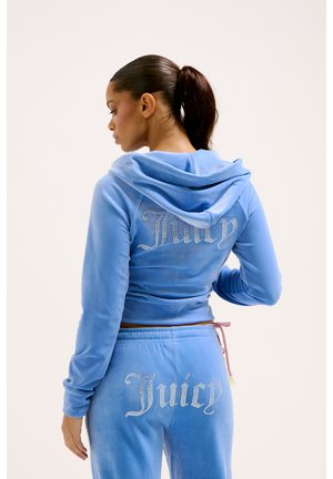 Tuta in velluto blu composto da una giacca cropped con cappuccio e pantaloni sportivi. Presenta decorazioni in strass "Juicy" sulla parte posteriore. Texture liscia.