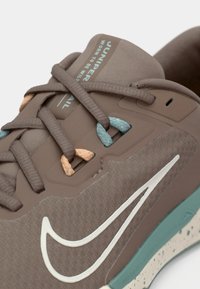 Gros plan sur une chaussure de course Nike marron avec des passants de lacets bleu sarcelle et orange, un logo swoosh blanc, et le texte "Juniper Trail Worn to Be Wild" sur la languette.