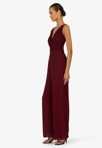 Burgunderfarbener Jumpsuit mit tiefem V-Ausschnitt, plissiertem Oberteil und weitem Bein. Aus weichem Stoff gefertigt, mit glatter Textur. Schwarze High-Heel-Sandalen.