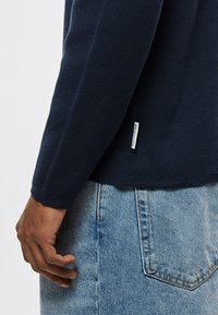 Tengerészkék kötött pulóver laza szabással, hosszú ujjakkal és oldalsó hasítékkal. Világoskék farmerrel, látható varrással párosítva.