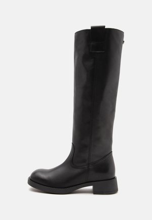 Botte haute en cuir noir avec un bout rond, un talon bloc bas et une languette en haut sur fond blanc.