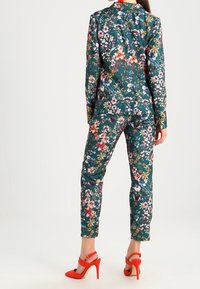 Floral suit made of fabric with a dark teal base. Features multicolored flowers, fitted design, and paired with red high-heeled sandals.