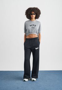 Sweatshirt cinza cropped com estampa "NIKE 1972", combinada com calças jogger pretas de corte largo com logótipo branco. O modelo usa óculos de sol escuros.