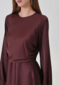Burgunderfarbenes Kleid mit langen Ärmeln und einem rundem Ausschnitt, gefertigt aus glattem Satinstoff mit gerafften Details an der Taille.