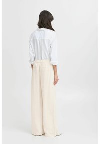 Camicia bianca con colletto indossata con pantaloni larghi beige. I pantaloni hanno una vita elastica e tasche laterali. Tessuto con una texture liscia.