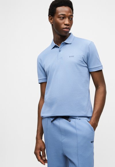Jeune homme portant un polo bleu clair et un pantalon assorti, les mains dans les poches, regardant sur le côté devant un fond uni.