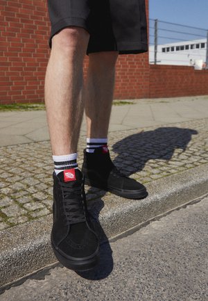 Czarne zamszowe sneakersy typu high-top o teksturowanej powierzchni i czarnych sznurówkach. Białe i czarne skarpetki w paski z widocznym czerwonym logo. Betonowa i brukowana nawierzchnia.