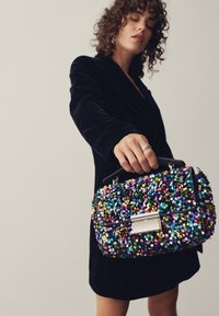 Daugiak spalvų blizgančių karoliukų rankinė su juodu rėmu, stačiakampio formos, blizgios tekstūros ir metaliniu užsegimu, kurią laiko juodą suknelę dėvinti žmogus.